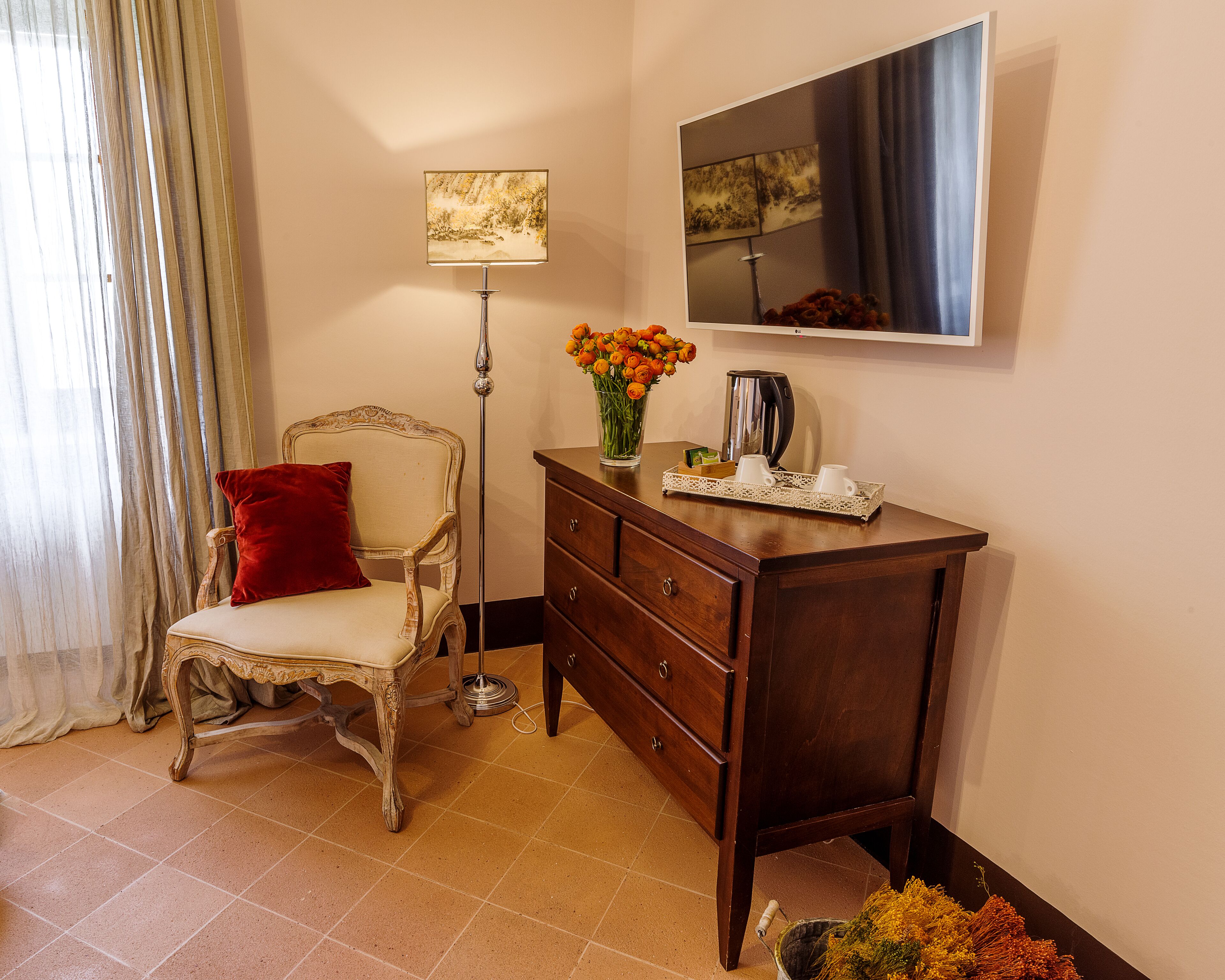 A room with a vintage chair, a wooden dresser, a vase of flowers, a floor lamp, and a painting on the wall.