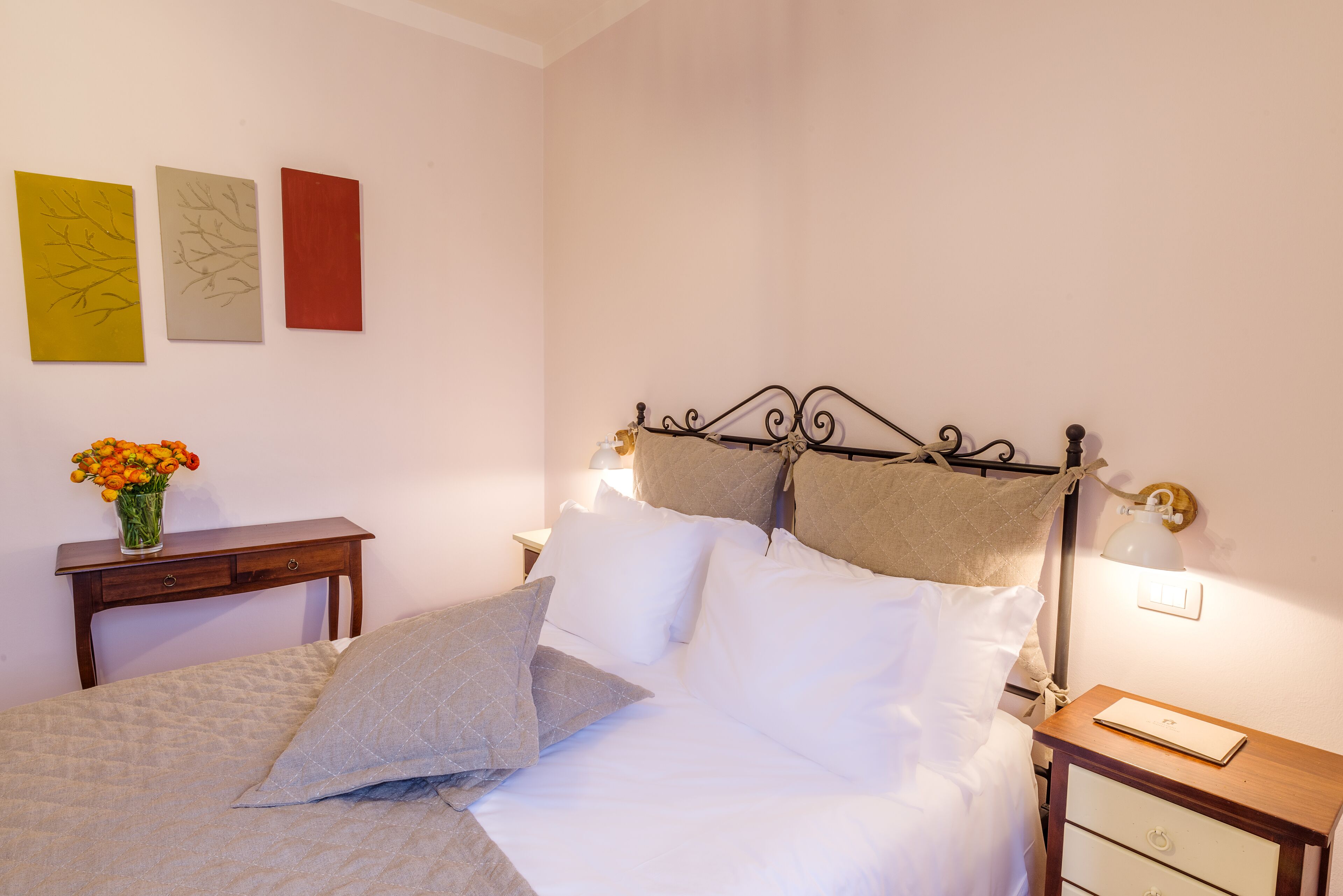 A neatly made bed with white linens and two pillows, a bedside table with a lamp, and a small nightstand with a lamp. There are three framed artworks on the wall.