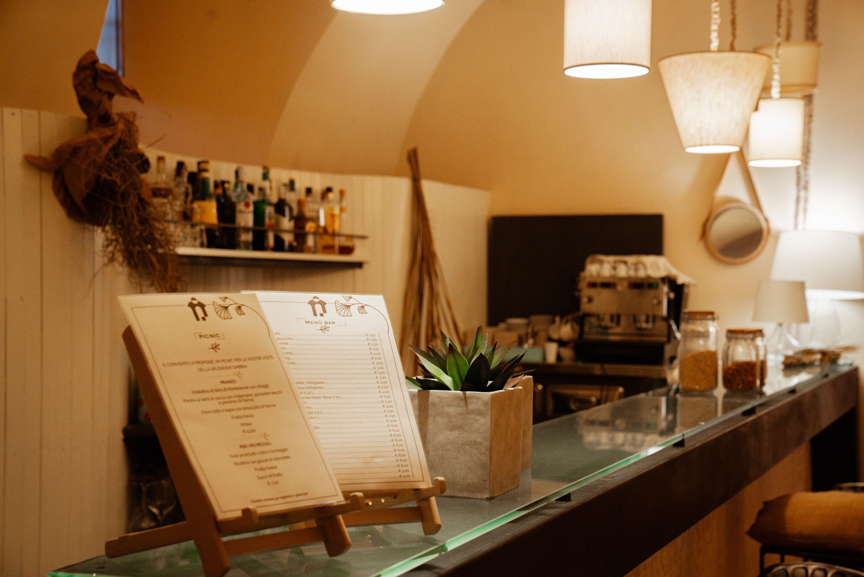 Zona de bar con una pizarra de menú, estantes llenos de botellas, una planta y una máquina de café.