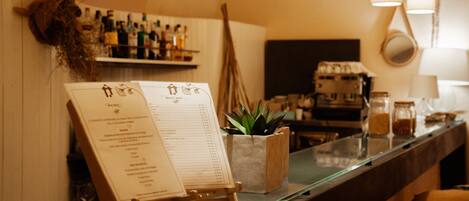 Zona de bar con una pizarra de menú, estantes llenos de botellas, una planta y una máquina de café.
