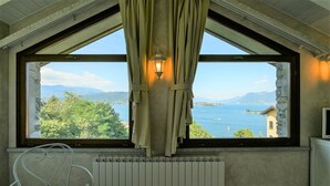 Een kamer met een groot raam met uitzicht op een meer en bergen, een radiator onder het raam en een metalen stoel aan de linkerkant.