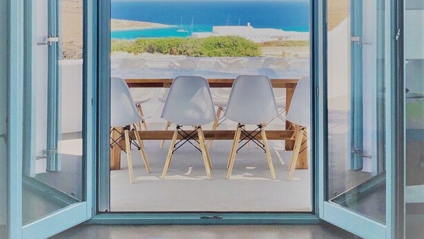 A room with a large glass door leading to a balcony with a view of the sea and boats. There are three white chairs on the balcony. - Blue Harpist Villas (Naxos)