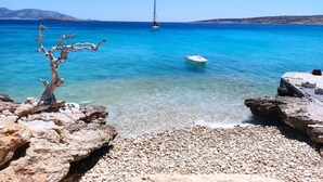 A rocky shoreline with clear blue water, a sailboat, and a tree in the foreground. - Blue Harpist Villas (Naxos)