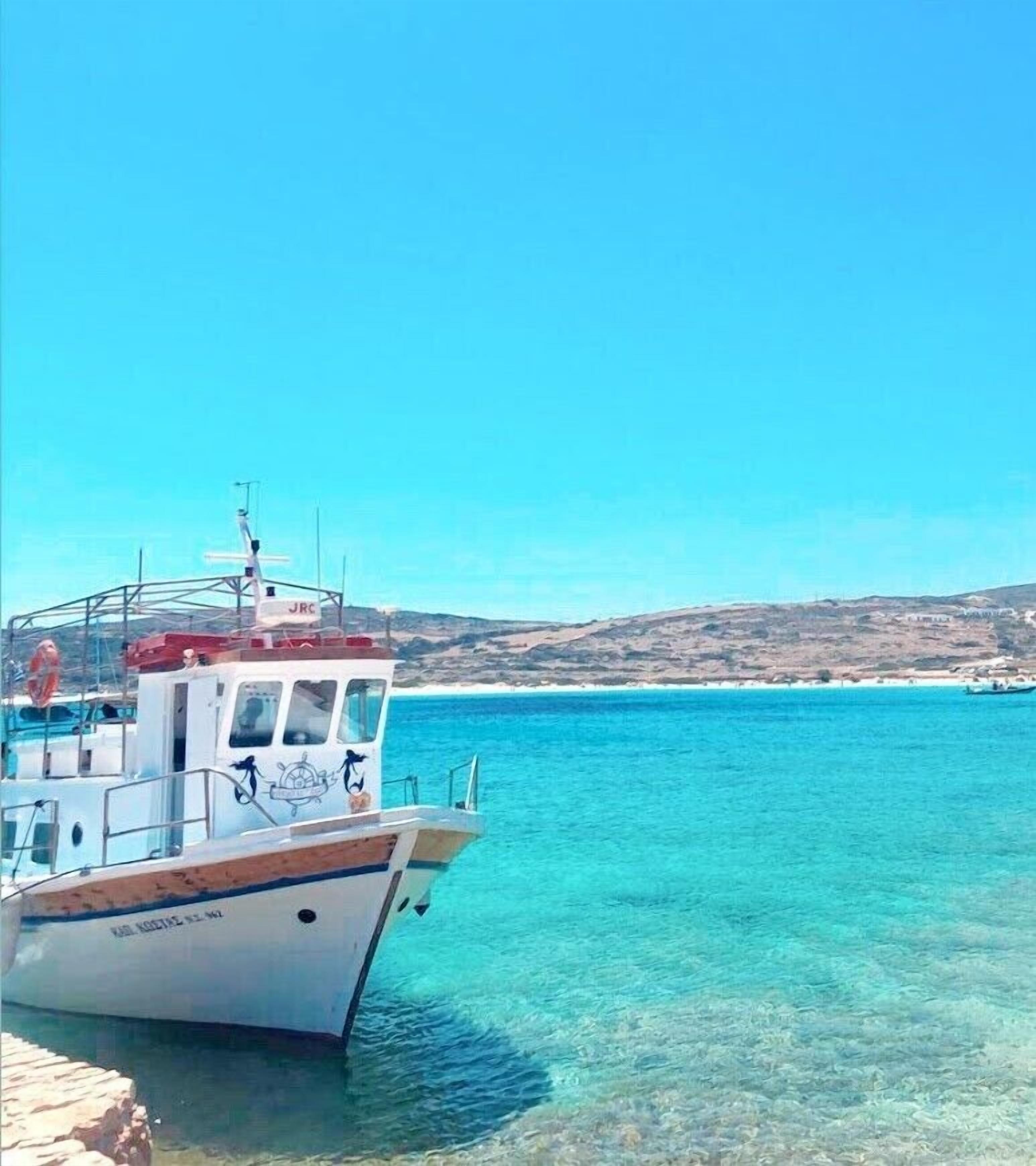 A boat docked at a pier with clear turquoise water and a hilly landscape in the background.