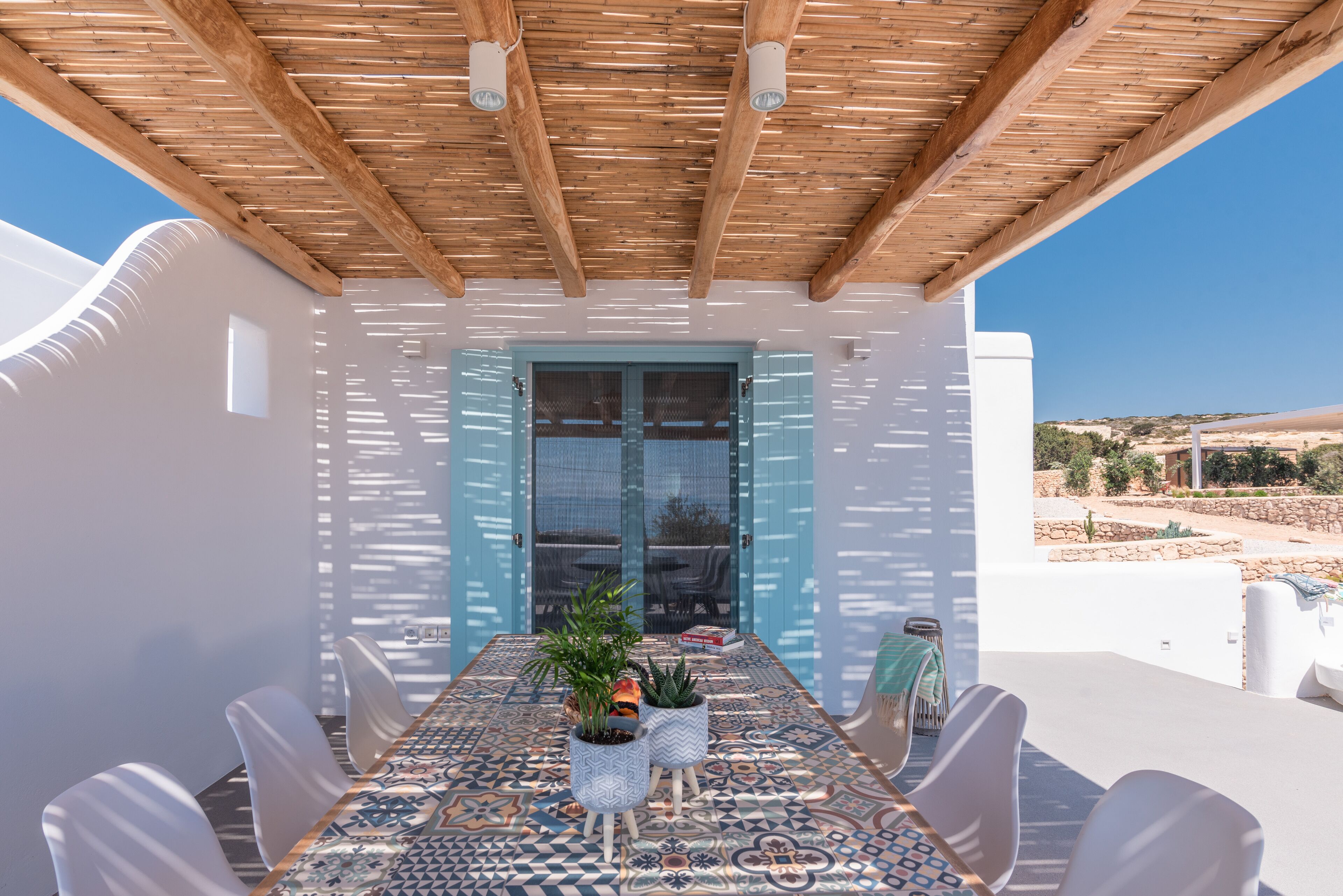 A covered outdoor dining area with a table set for six, surrounded by white chairs, and a view of the landscape.