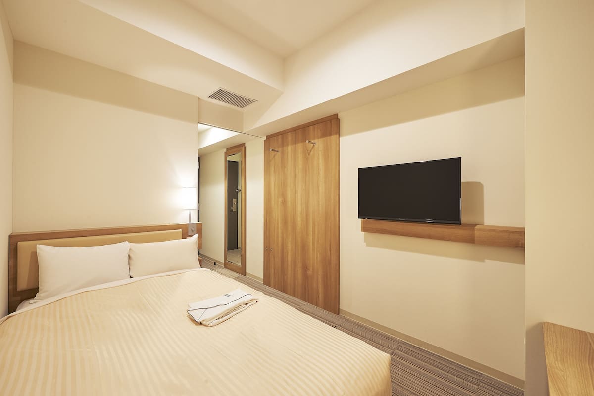 A hotel room with a bed, a television mounted on the wall, and a wooden wardrobe.