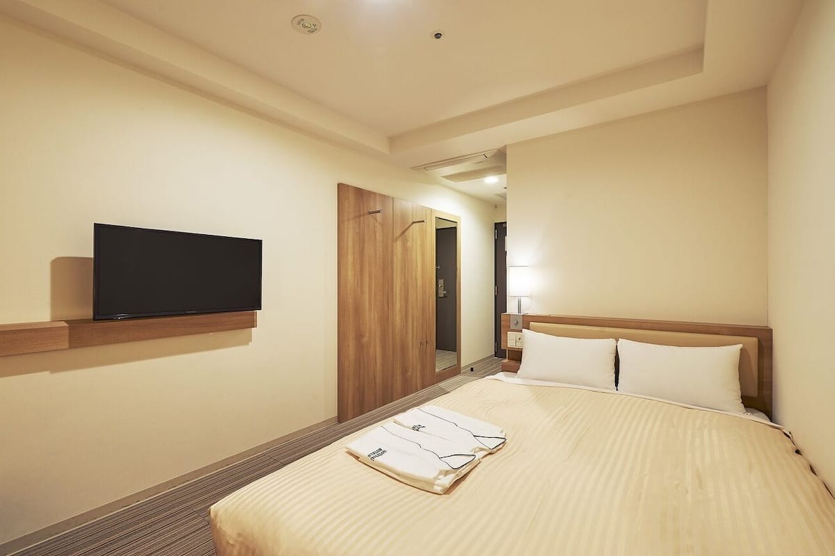 A hotel room with a bed, a television mounted on the wall, and a wooden wardrobe.