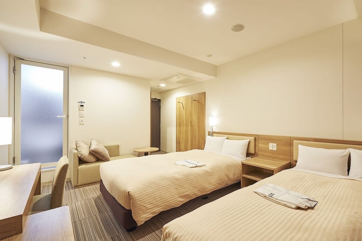 A hotel room with two beds, a desk, a chair, a small table, and a door leading to a bathroom.