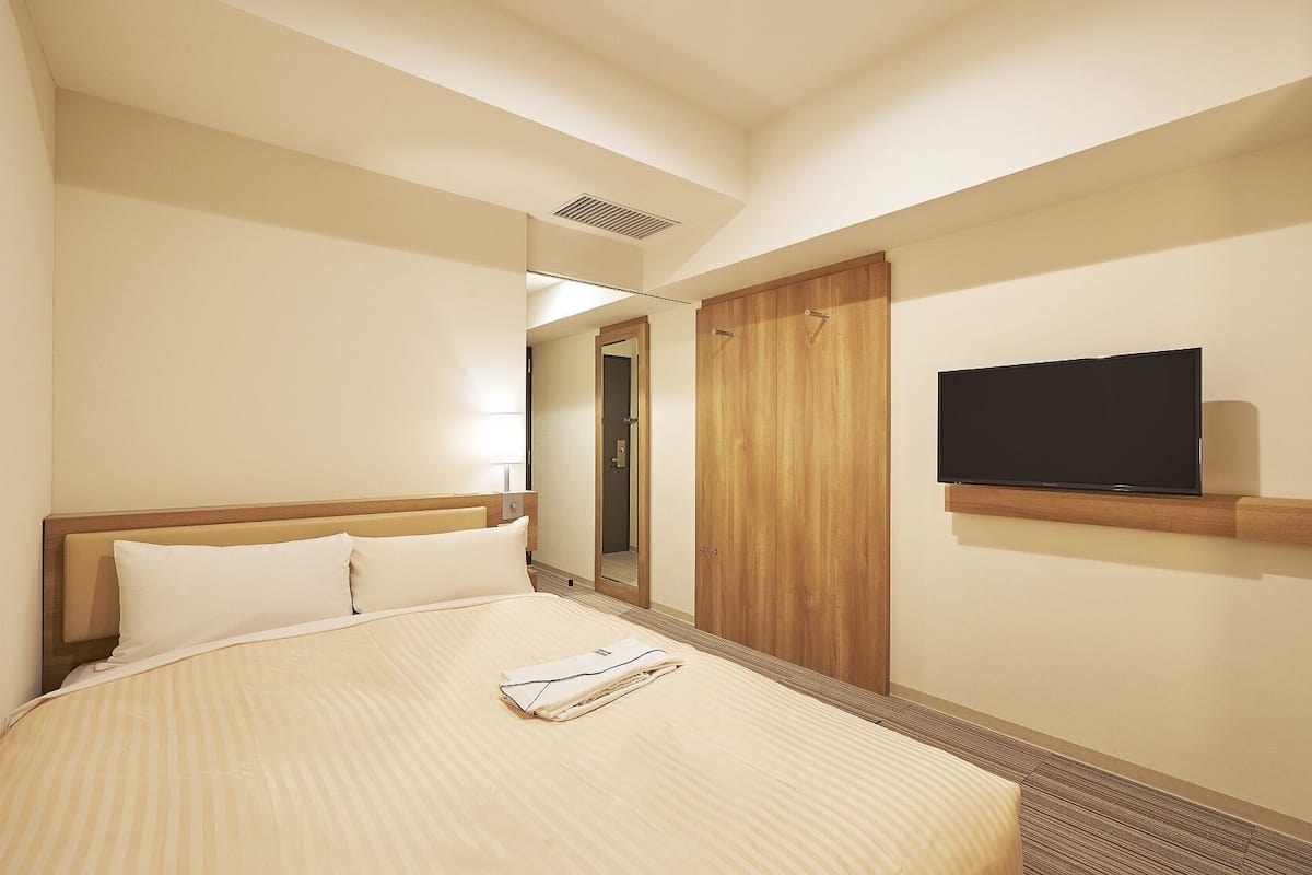 A hotel room with a bed, a TV mounted on the wall, and a wooden wardrobe.