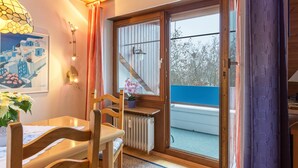 Quarto com mesa de madeira, cadeiras, radiador, janela com vista para árvores e porta que dá para o exterior.