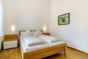 A bedroom with a wooden bed, white bedding, a nightstand, a drawer, and a wall-mounted painting.