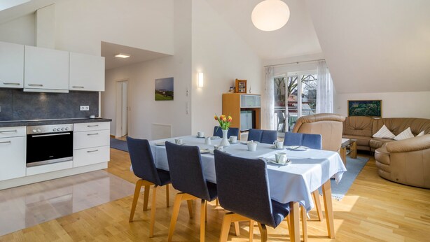 Una cocina moderna con armarios blancos, una mesa de comedor para cuatro personas y una sala de estar con sofá y un cuadro.