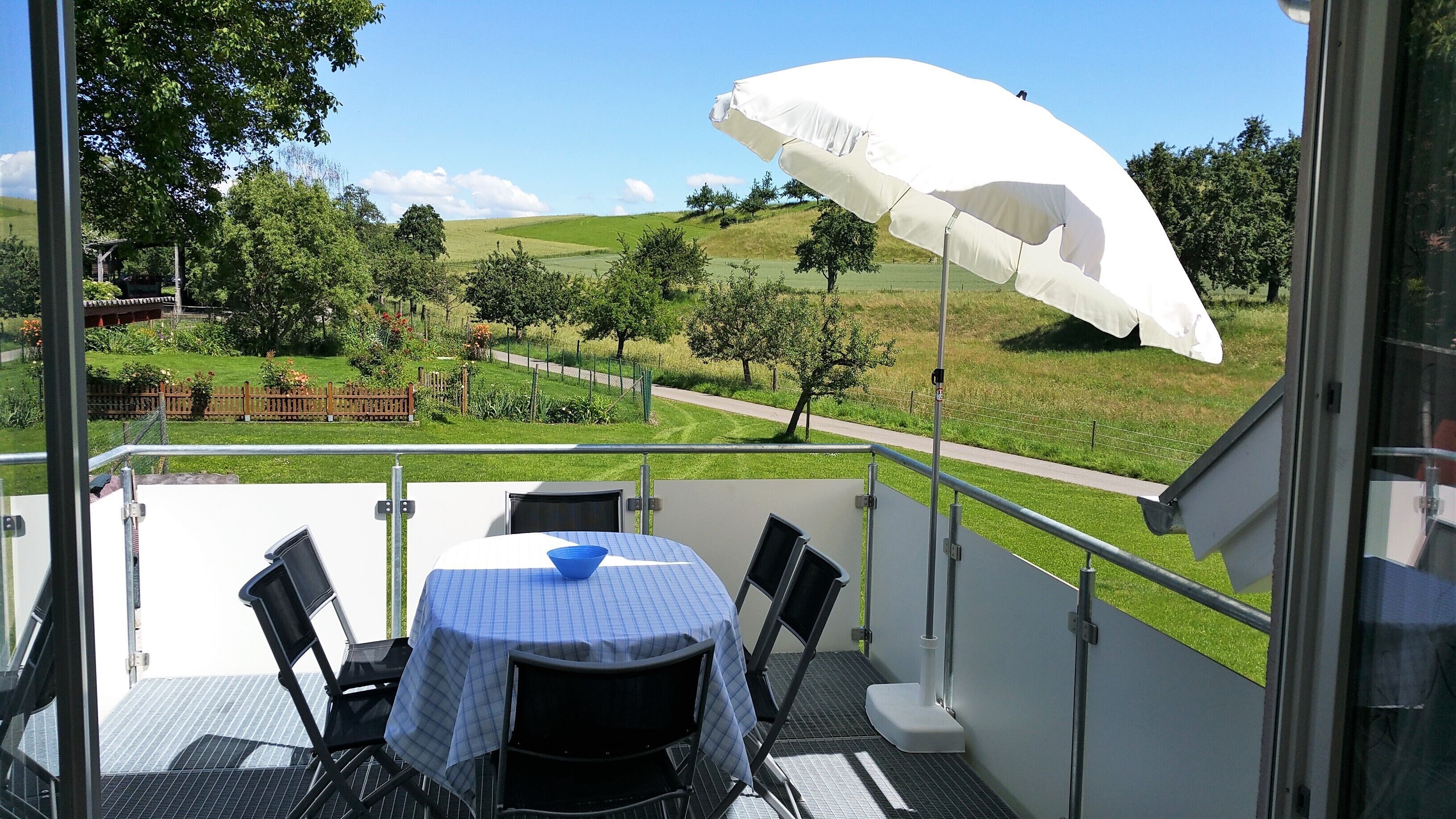 A balcony with a table and chairs, a white umbrella, and a view of a green landscape with trees and a path.