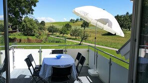 A balcony with a table and chairs, a white umbrella, and a view of a green landscape with trees and a path.