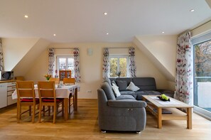 A modern living room with a sofa, dining table, and chairs, a kitchen area with cabinets, and large windows with curtains.