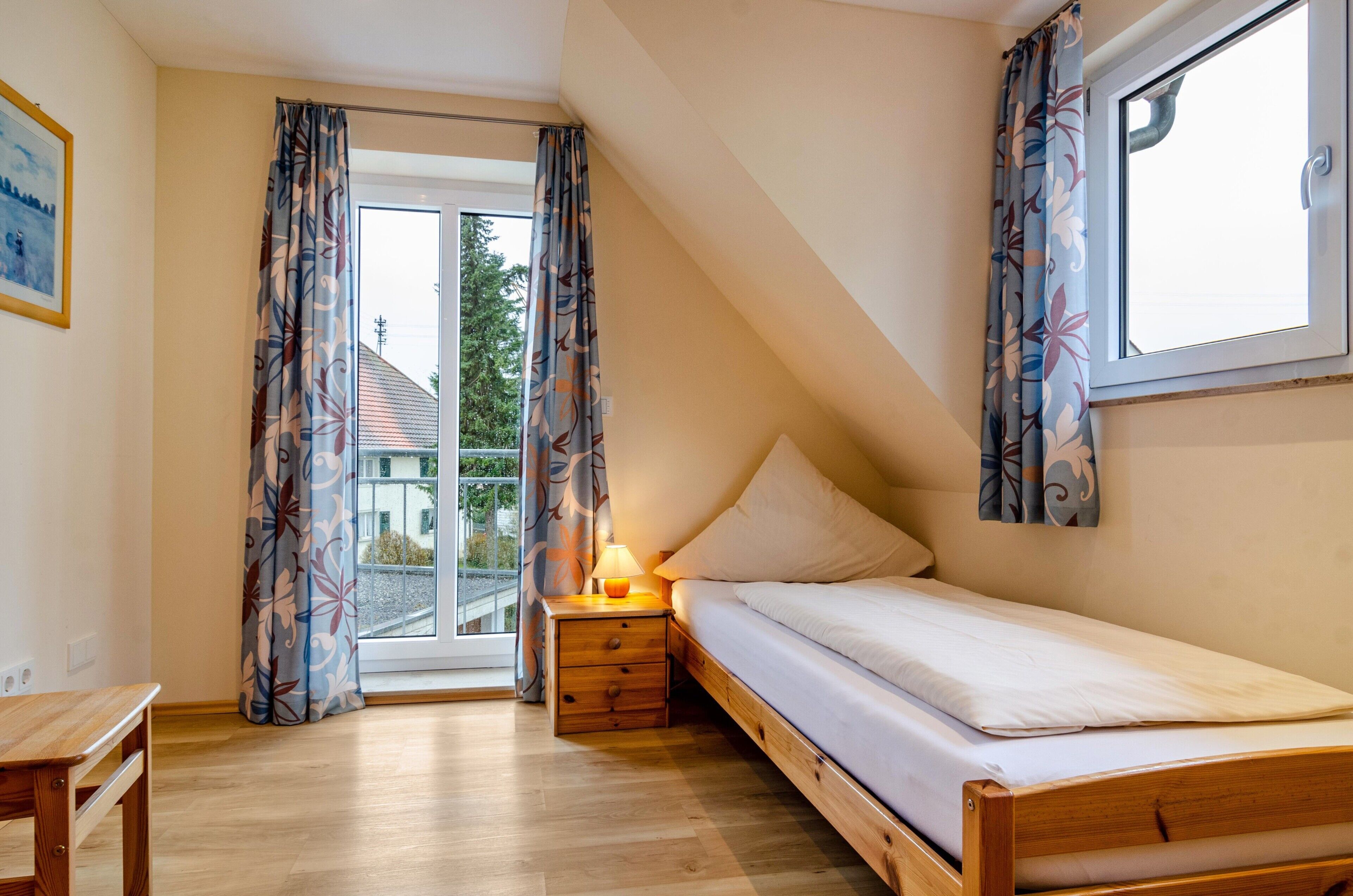 A bedroom with a bed, a nightstand with a lamp, a window, and a door leading to a balcony.