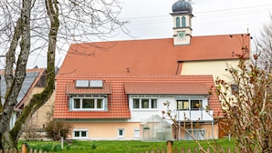 Ein zweistöckiges Haus mit rotem Ziegeldach, einem sichtbaren Schornstein und einem kleinen Anbauten Gewächshaus.