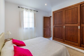A bedroom with a bed, a nightstand, a lamp, a window with curtains, and a wooden wardrobe.