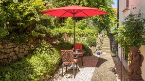 Um pátio de jardim com um guarda-sol vermelho, mesa e cadeiras, rodeado por vegetação exuberante e uma parede de pedra.