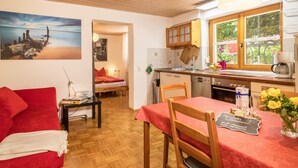 Um quarto aconchegante com um sofá vermelho, uma mesa de jantar com toalha vermelha, uma área de cozinha com armários de madeira e uma grande janela com vista para o verde.