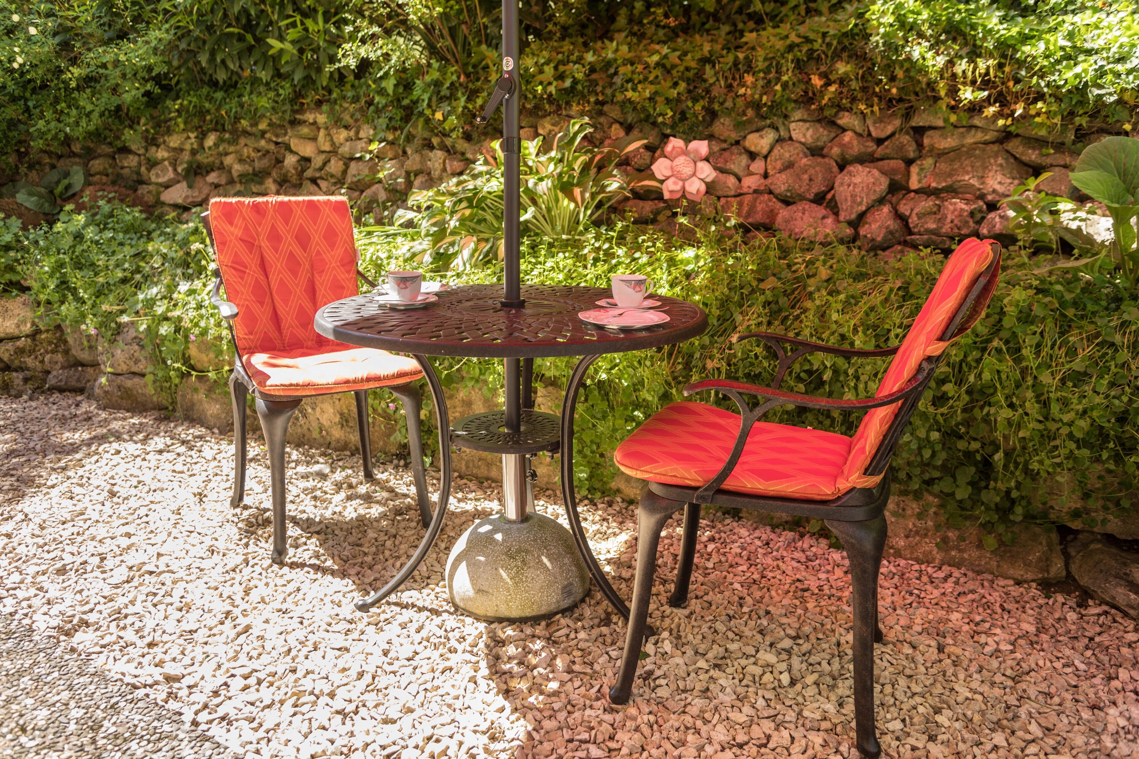 A round metal table with two red cushioned chairs, a stone base, and a backdrop of greenery and a stone wall.