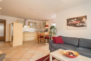 A modern living room with a grey sofa, a dining area with a wooden table and chairs, and a kitchen with light-colored cabinets and a red pendant light.