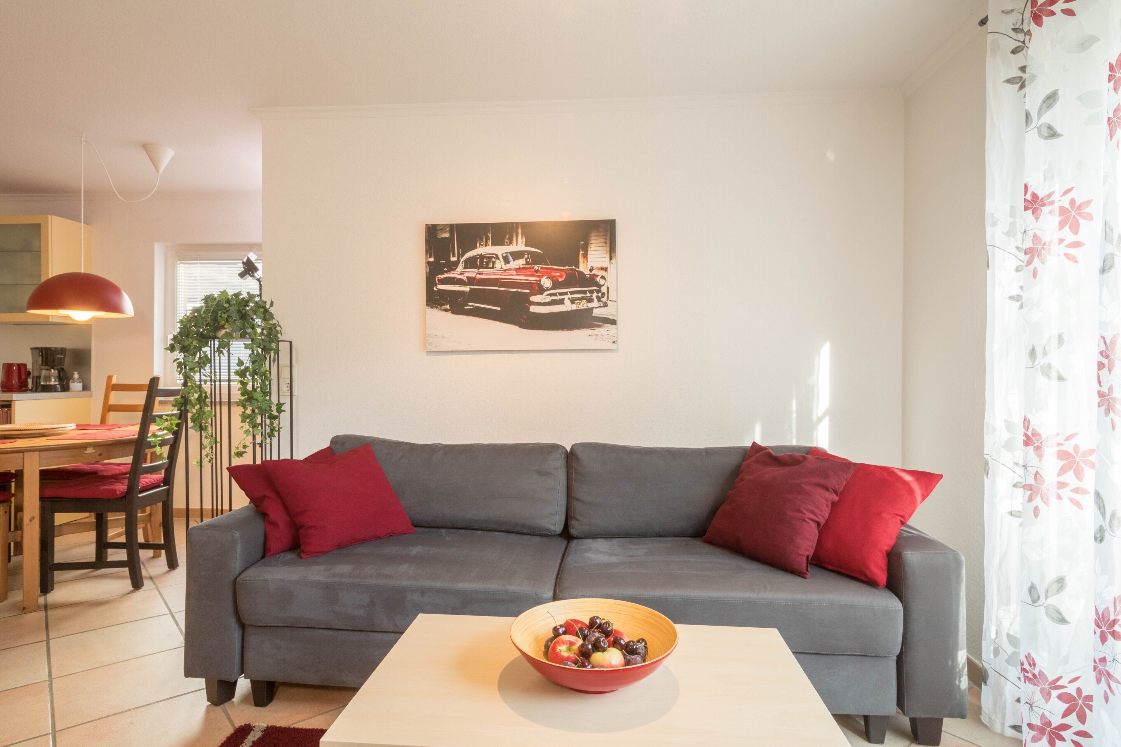 Ein Wohnzimmer mit grauem Sofa, roten Kissen, einer Obstschale auf einem Tisch, einem Essbereich mit einem roten Stuhl und einem an der Wand hängenden Bild von einem Auto.