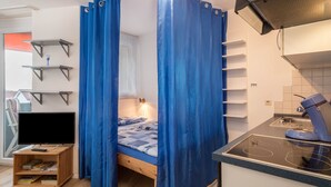 A room with a bed, a TV on a wooden stand, a kitchen area with a stove, and a window with curtains. - Apartment “Förster” with Balcony and Wi-Fi (Überlingen)