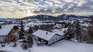 Paisatge cobert de neu amb muntanyes al fons i una casa amb teulada blanca al primer terme.