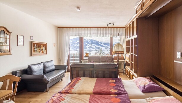 Habitació amb un sofà, un llit amb llençol estampat, un armari de fusta i vista a les muntanyes per la finestra.
