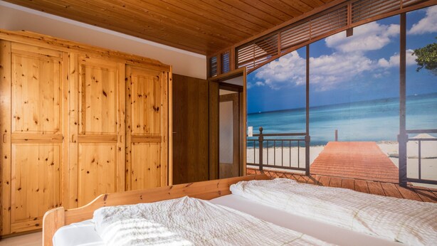 A wooden room with a bed, a wardrobe, and a large window with a beach view. - Holiday Apartment "Monika Haug" with Lake View, Mountain View, Wi-Fi, Balcony & Garden (Immenstaad am Bodensee)