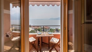 Een balkon met een tafel en stoelen, uitzicht op zee, en een gebouw op de achtergrond.