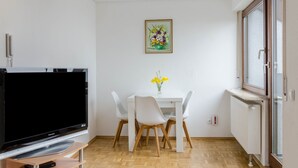 A small room with a TV, white chairs, a table with a vase of flowers, and a window.