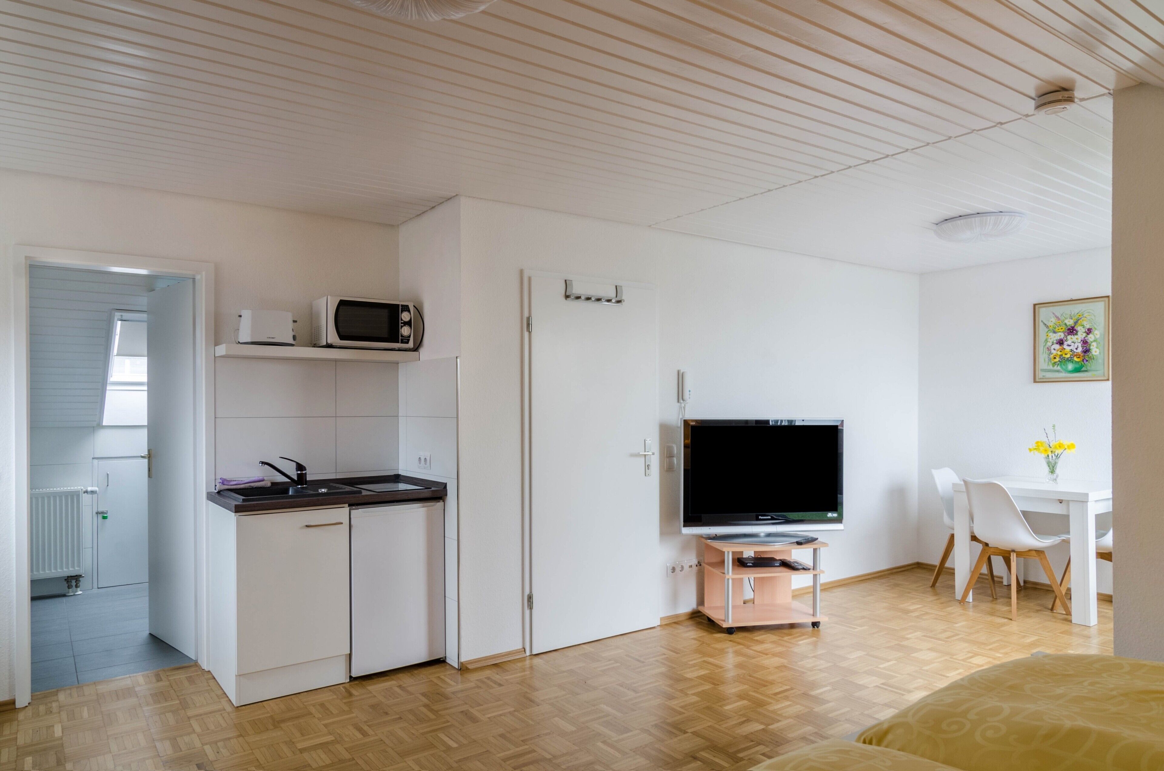 Ein kompaktes Zimmer mit Küchenzeile, einem Flachbildfernseher, einer Essecke mit Tisch und Stühlen sowie einem Bett.