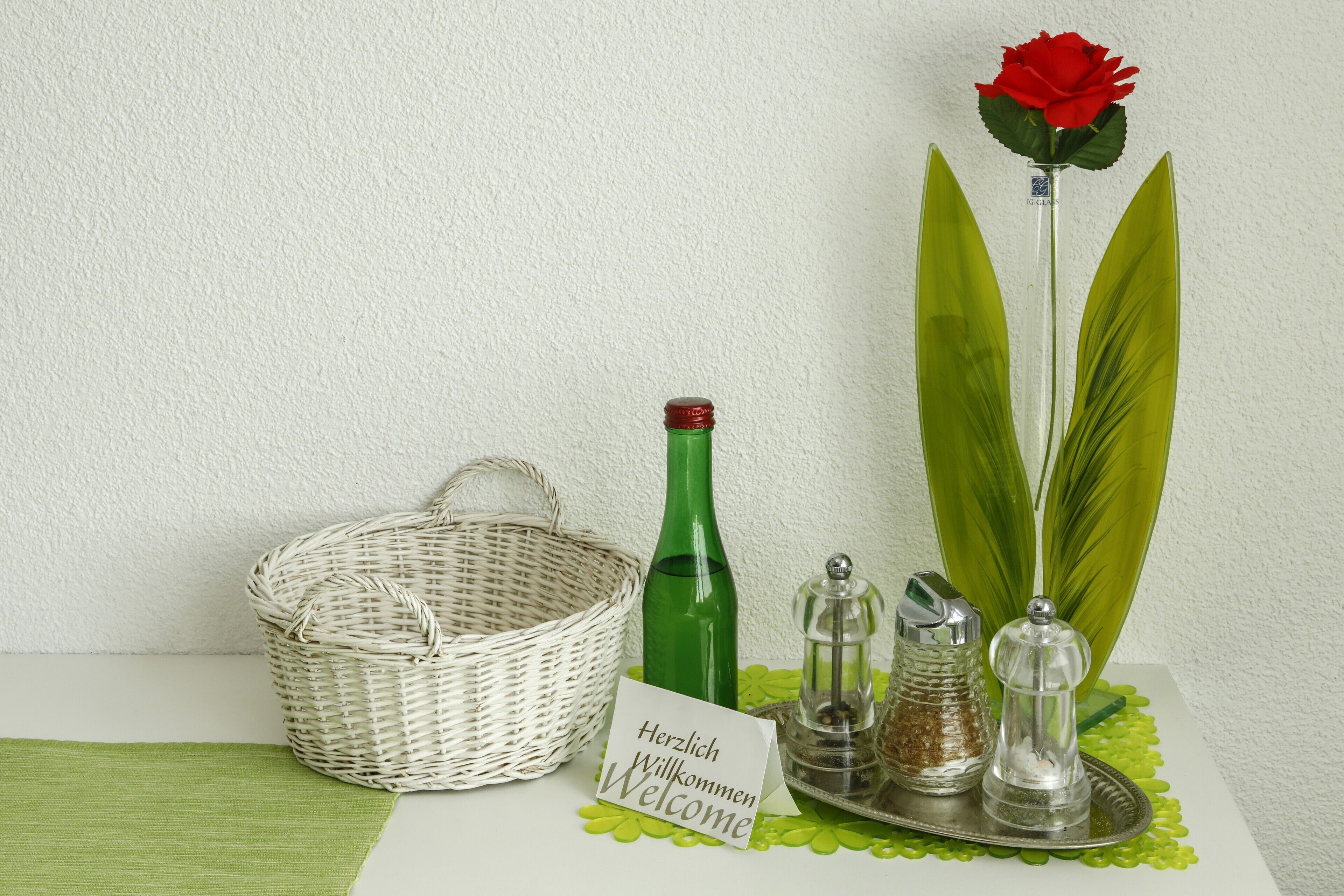 Ein Willkommensschild, eine rote Rose, eine grüne Flasche, ein Salzlöffel, ein Pfefferlöffel und ein Korbwägelchen auf einer Tischplatte.