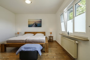 Une chambre à coucher avec un lit en bois, un banc, une table de chevet, une lampe fixée au mur, un radiateur et une fenêtre avec des stores.