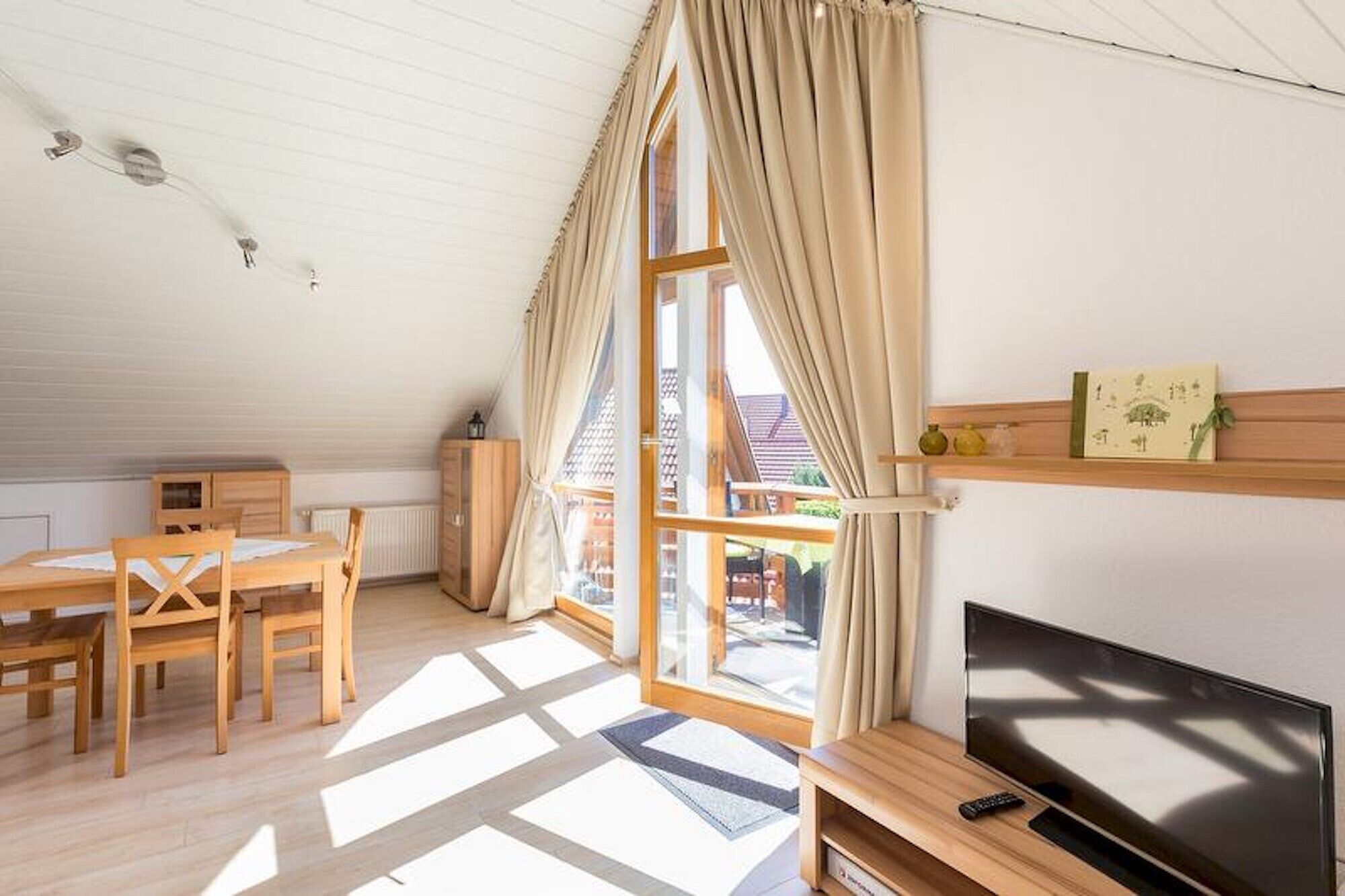 Una habitación con una mesa de comedor de madera, sillas, un televisor sobre un mueble de madera y ventanas grandes con cortinas.