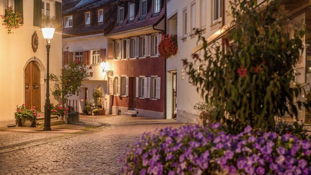 En kullerstensgata med traditionella europeiska byggnader, en gatlykta och blomlådor.
