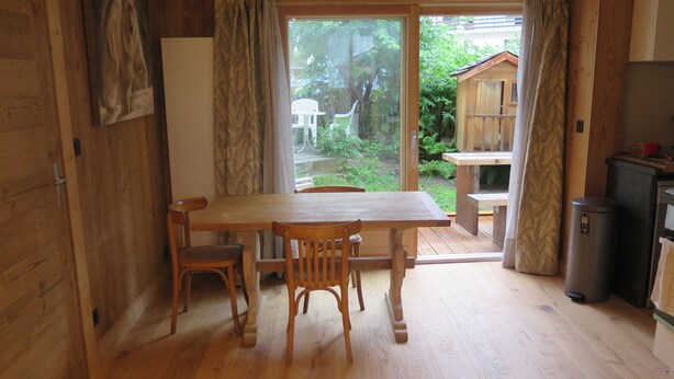A wooden interior with a dining table and chairs, a refrigerator, and a sliding glass door leading to an outdoor area with a wooden deck and greenery.