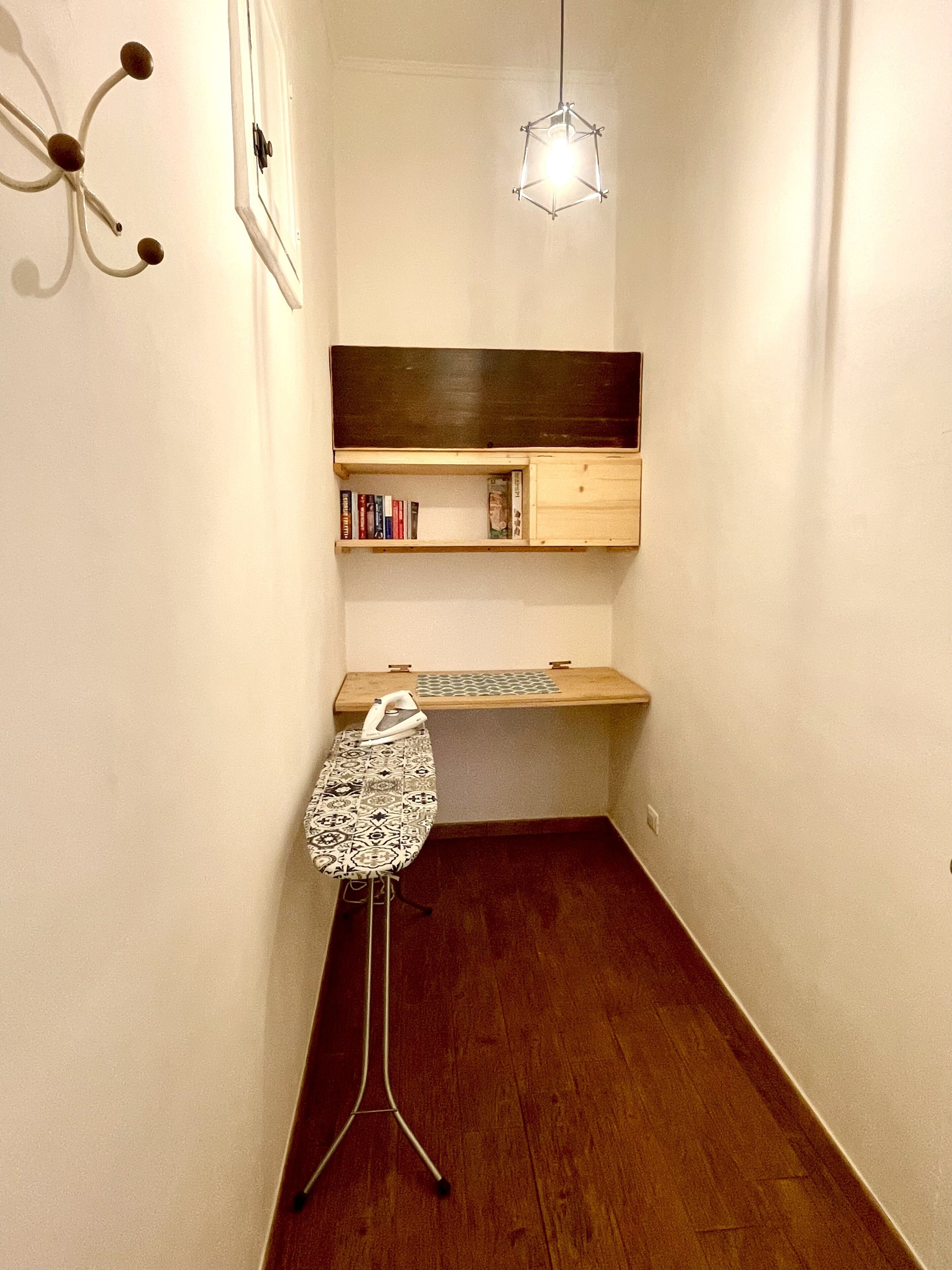 A narrow hallway with a wooden floor, a small ironing station, a wall-mounted shelf with books, and a hanging light fixture.