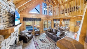 A spacious log cabin interior with a stone fireplace, leather furniture, a balcony, and a large TV screen. - A Higher Calling, Spacious Smoky Mountain Getaway –Perfect for Families & Groups (Pigeon Forge)