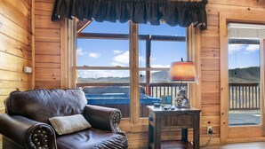 A wooden cabin interior with a leather couch, a small table with a lamp, and a view of mountains through a large window. - A Higher Calling, Spacious Smoky Mountain Getaway –Perfect for Families & Groups (Pigeon Forge)