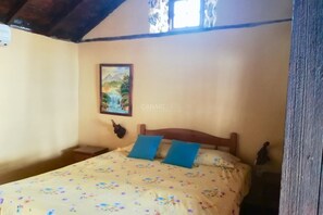 A bedroom with a wooden bed, floral bedspread, two blue pillows, a wall-mounted painting, and a small decorative item on the nightstand.