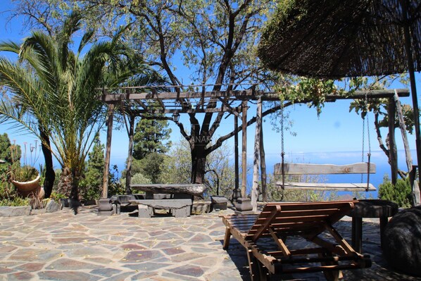 Un patio con suelo de piedra, sillones de madera, una hamaca y una pérgola con plantas colgantes.
