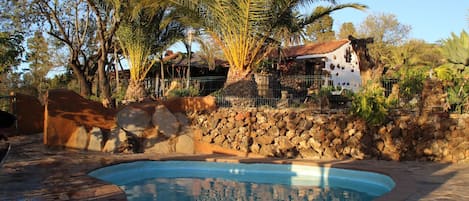 Una piscina circondata da un muro in pietra, palme e un edificio con facciata bianca.