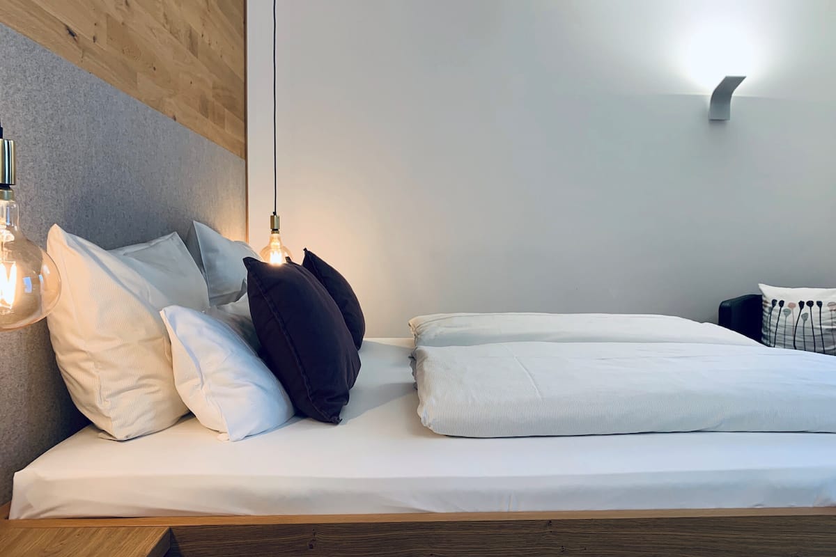 Un lit bien fait, avec du linge de lit blanc et plusieurs oreillers, une tête de lit en bois et un luminaire fixé au mur.