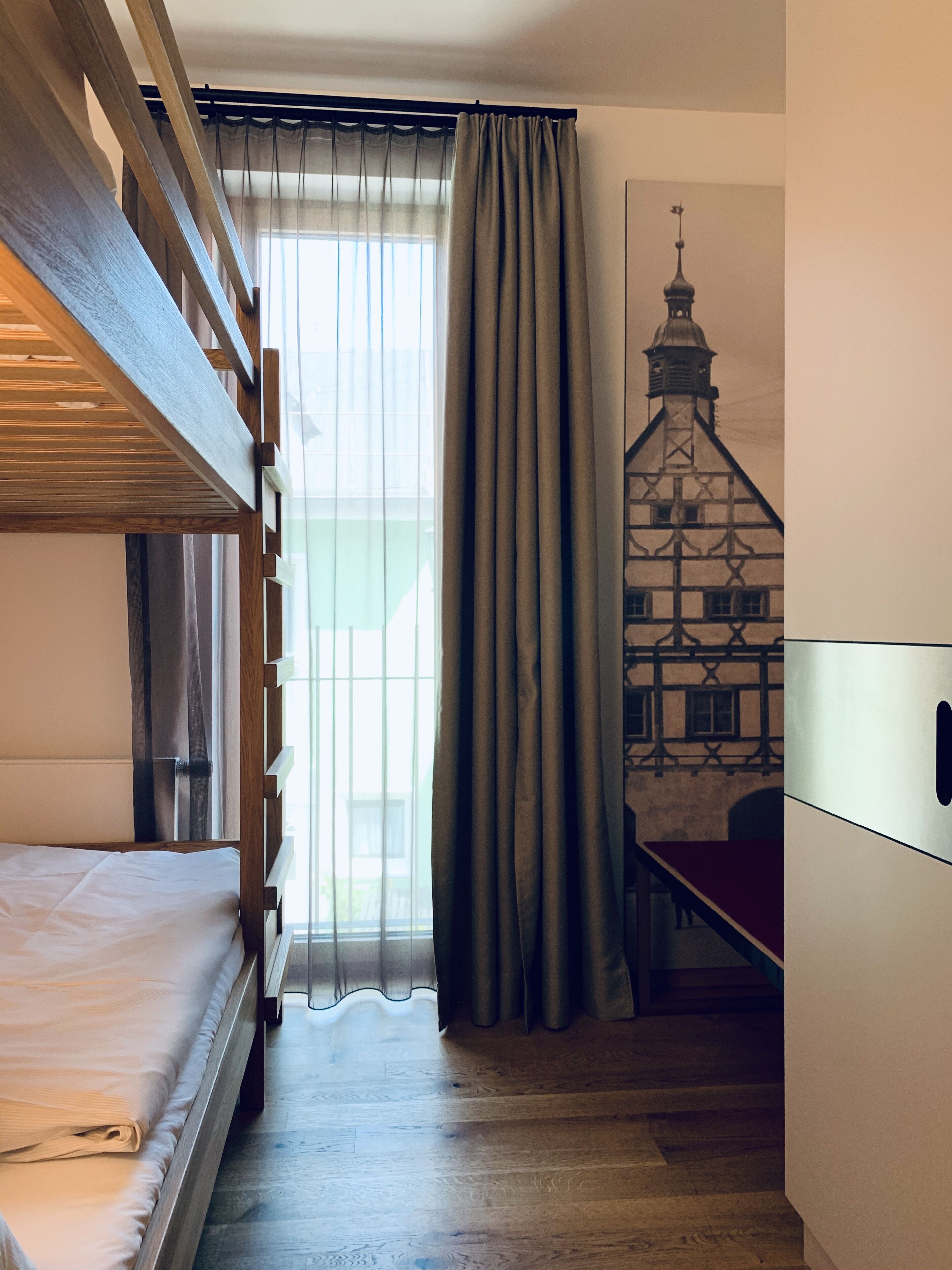Een kamer met een stapelbed, een raam met dunne gordijnen, uitzicht op een gebouw met een toren, en een trap naar een hoger gelegen niveau.