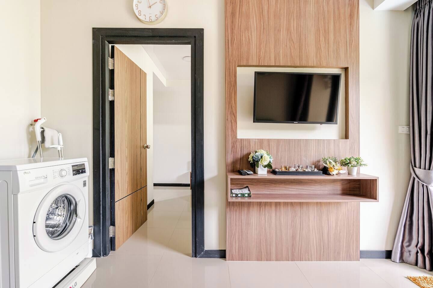 A modern living room with a flat-screen TV mounted on a wooden wall, a washing machine, and a small shelf with decorative items.