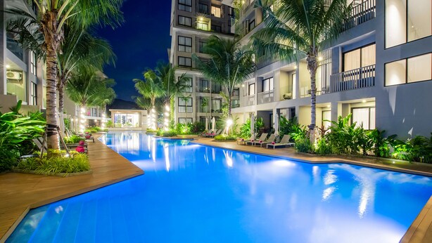 A swimming pool with lounge chairs and palm trees, surrounded by a multi-story building with balconies.