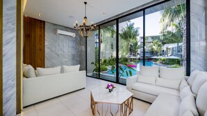 A modern living room with a white sofa, a round coffee table, and a large window offering a view of a swimming pool and greenery.